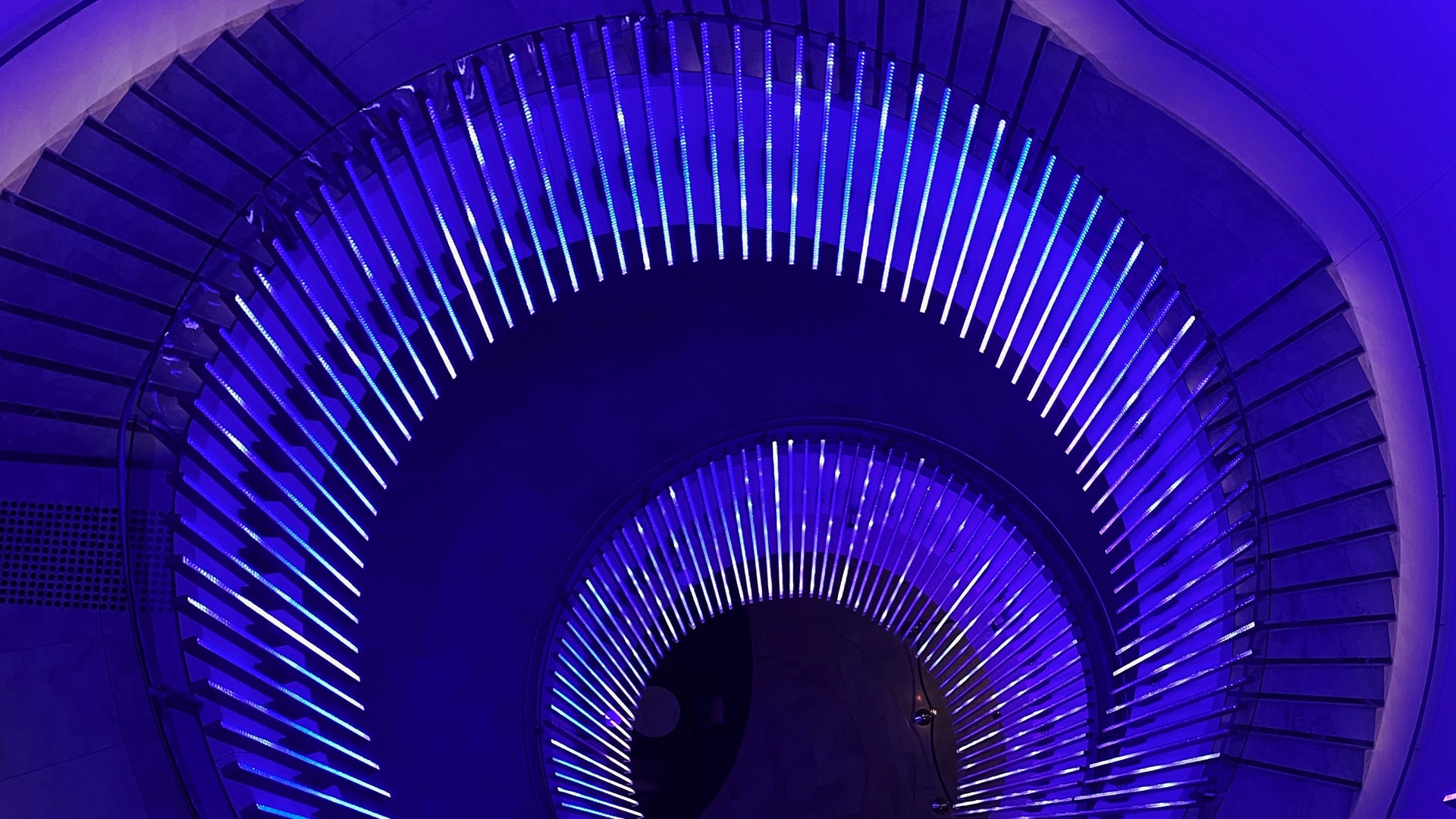 Spiralling staircase railings at night, illuminated in neon blue light. 